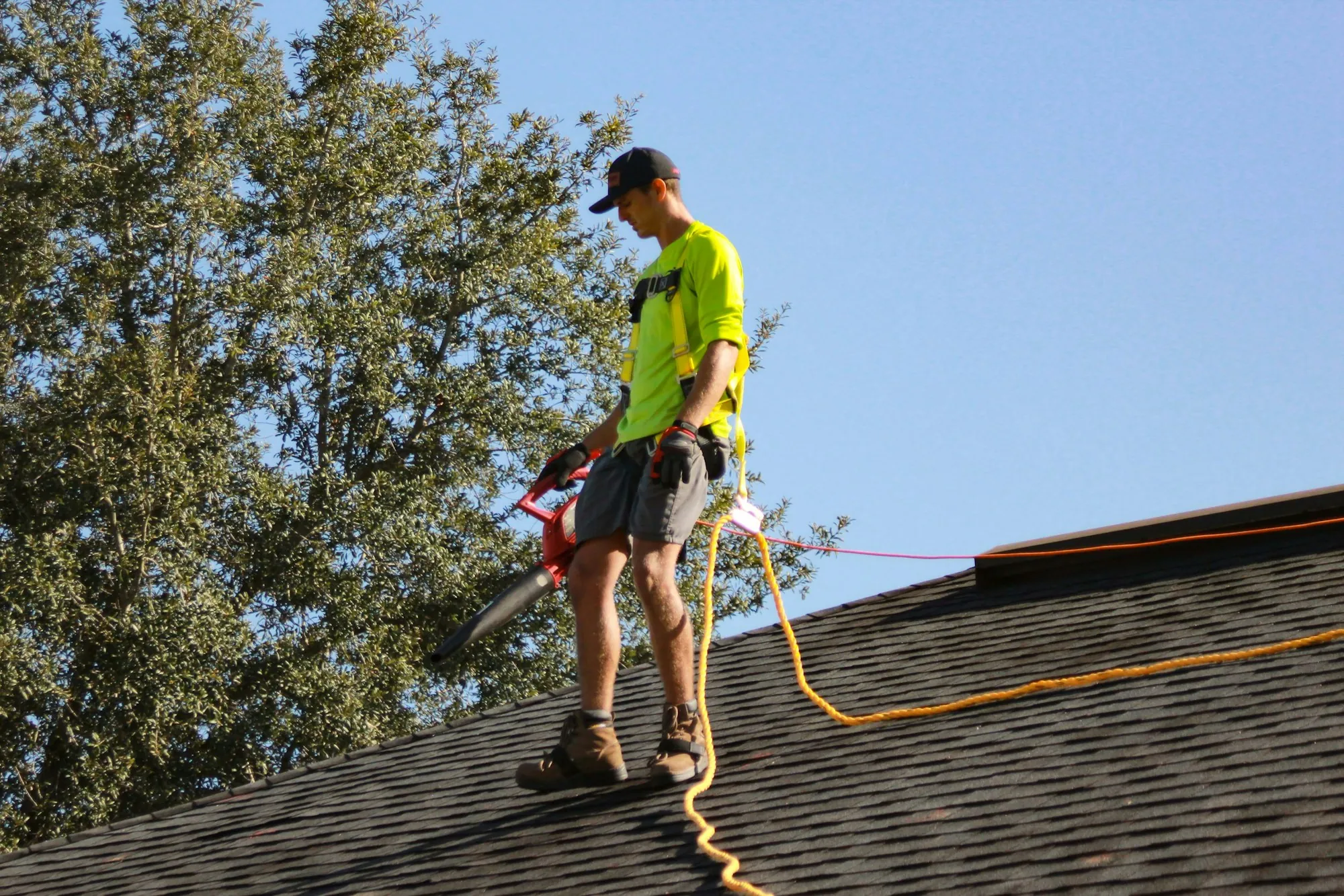 Roofer in a hi-vis shirt and harness standing on a shingle roof near trees